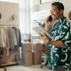 Mann telefoniert und nutzt ein Tablet in einem Mode-Showroom mit Kleidung, Kartons und Arbeitsbereich.
