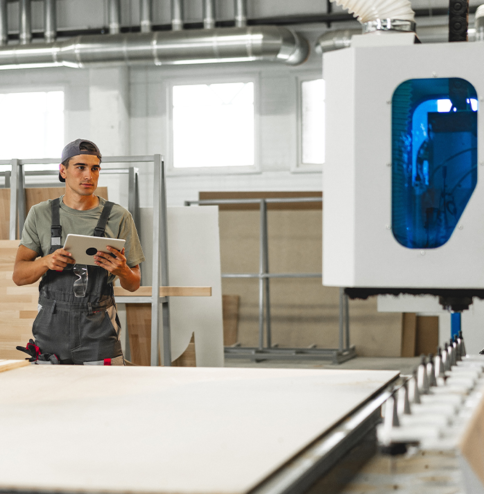 Worker in a cap and overalls using a tablet to monitor a machine in a furniture manufacturing facility